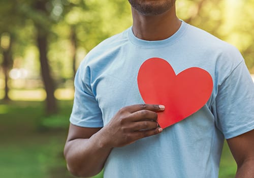 a person in a blue t shirt standing outside holds a red paper heart against their chest